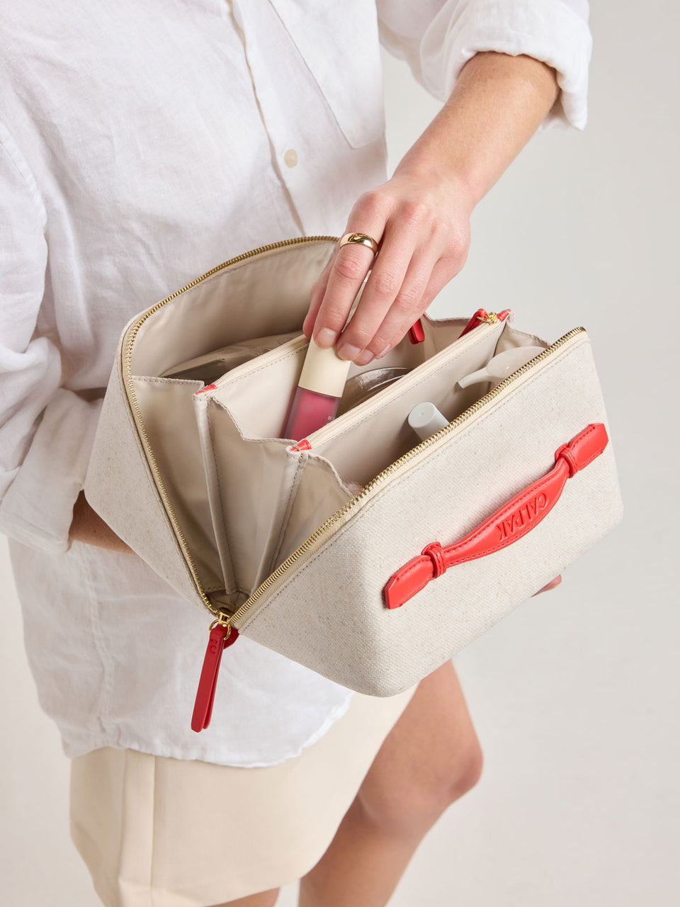 Model putting item into CALPAK Colette Lay-Flat Canvas Toiletry Bag in beige canvas material and red finishing; CLTB2601-NATURAL-TOMATO