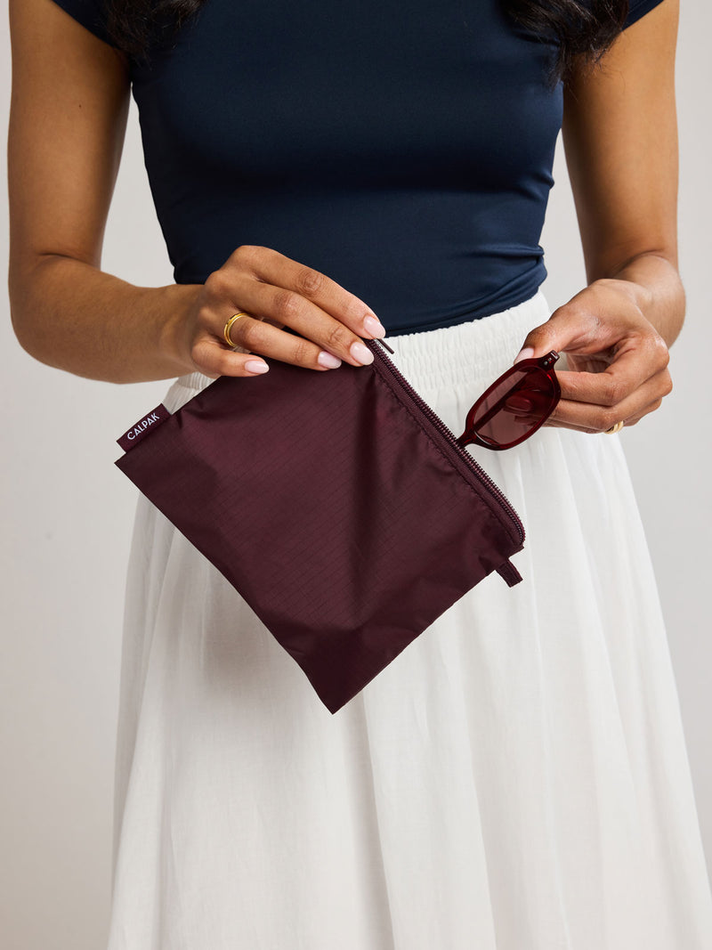 A woman in a navy top and white skirt holds a burgundy Dark Cherry CALPAK Compakt Zippered pouches of different sizes, arranged in a fan-like layout on a clean white background