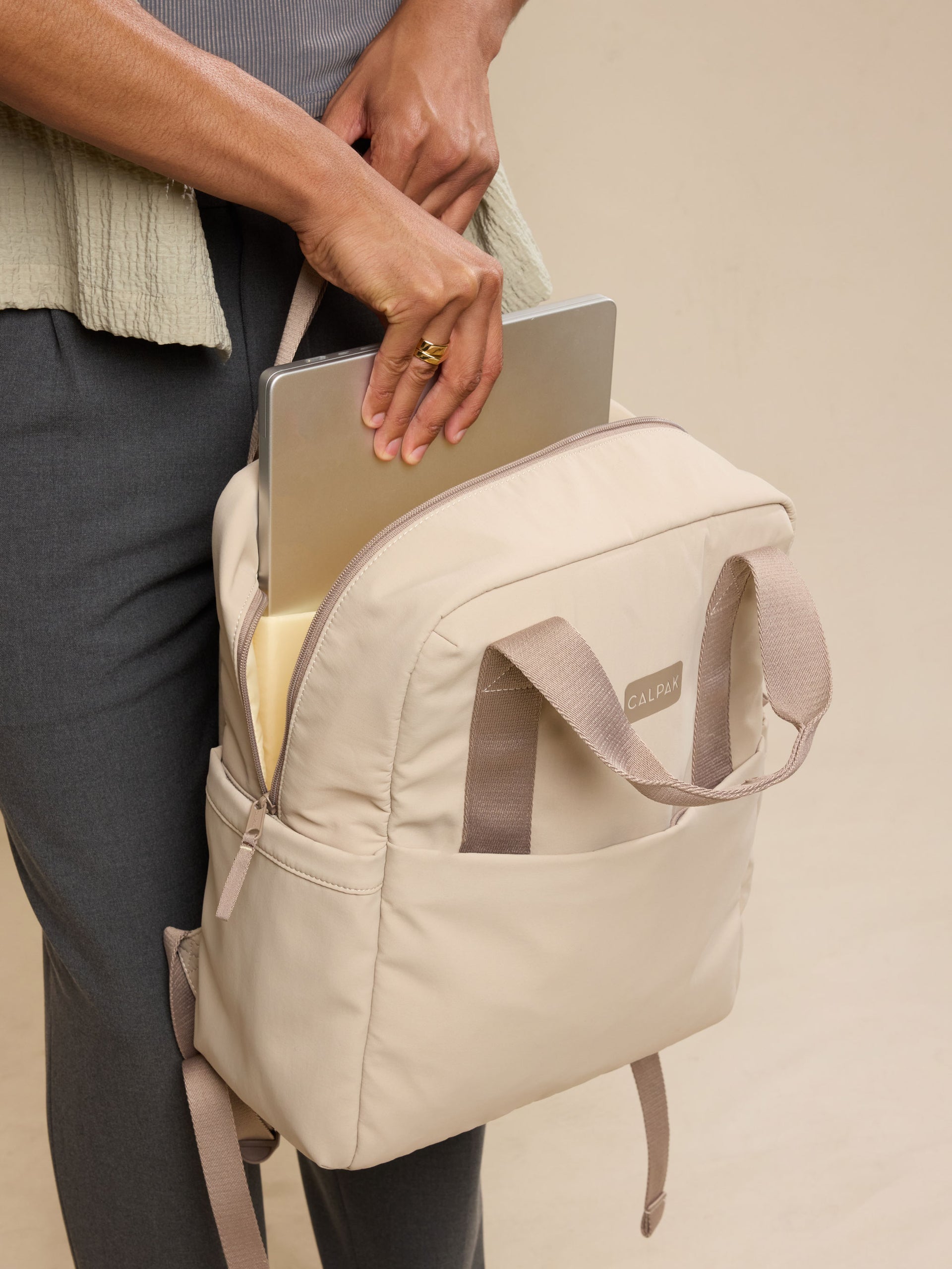 CALPAK Connect Laptop Backpack in grey beige Greige with model putting in laptop into backpack; BCN2401-GREIGE
