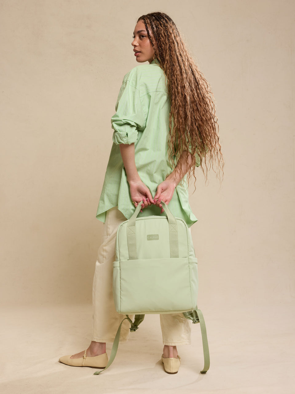 CALPAK Connect Laptop Backpack in light green Meadow being held by model front view on a beige background; BCN2401-MEADOW