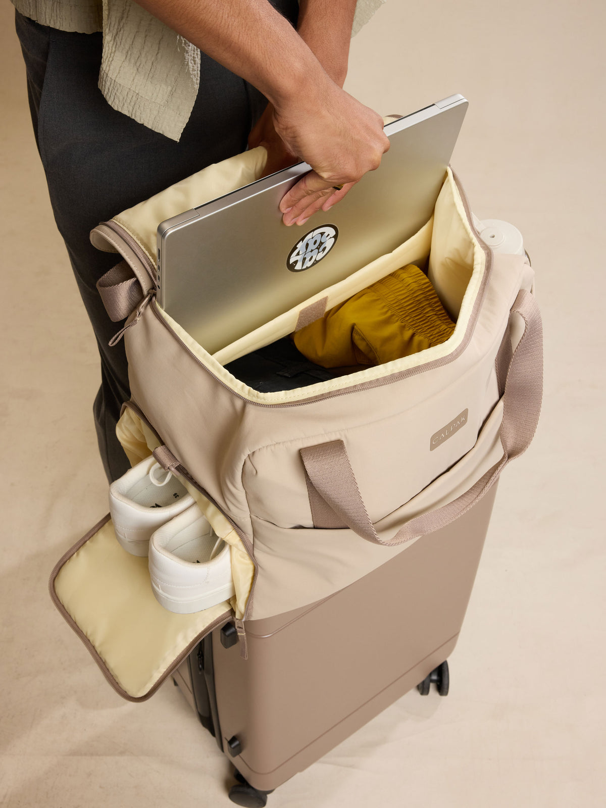 CALPAK Laptop Duffel in beige Greige with model putting in laptop in duffel against a beige background; DCN2401-GREIGE