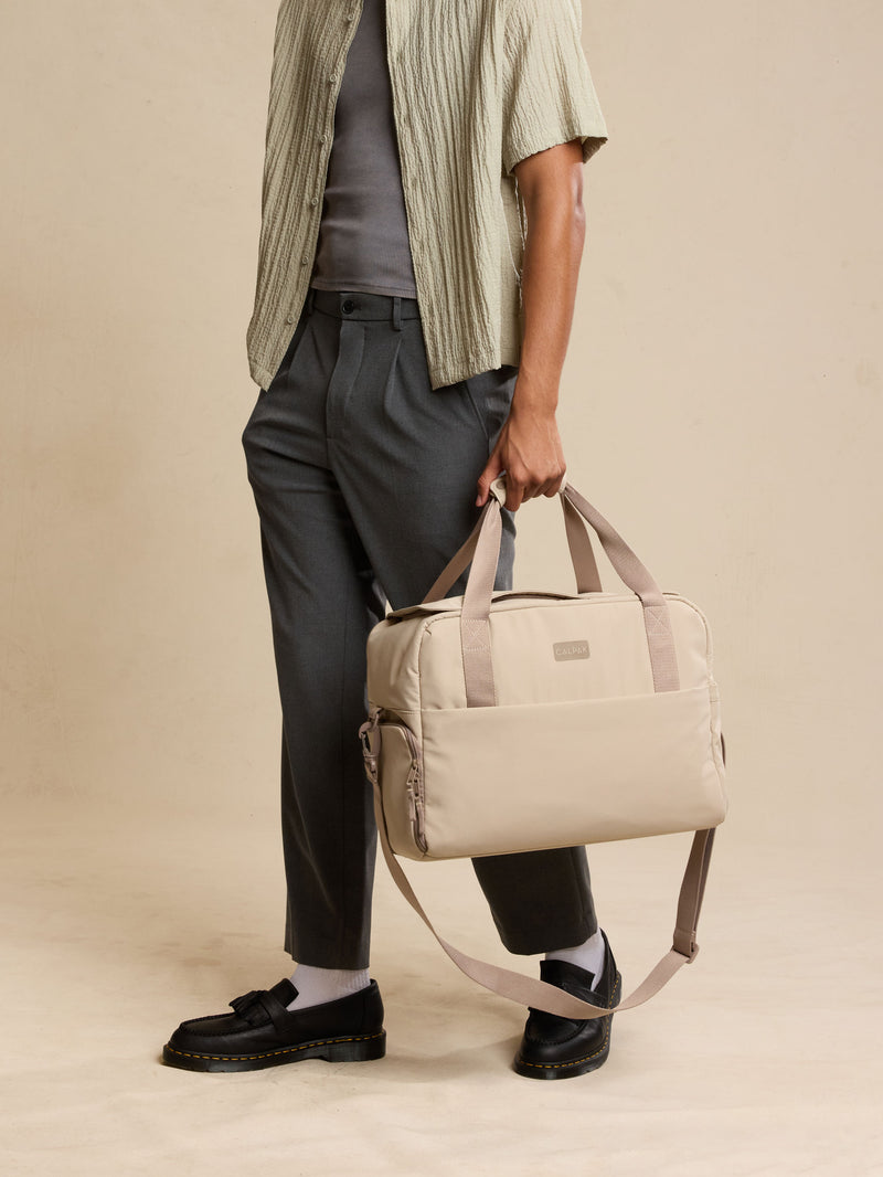 CALPAK Laptop Duffel in beige Greige being held by model with beige background