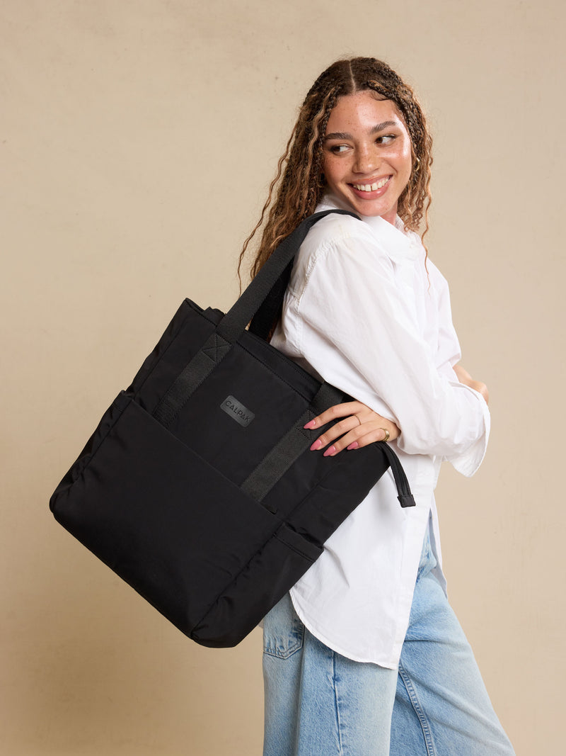 CALPAK Zippered Laptop Tote Bag in Black  on a beige background being worn by model