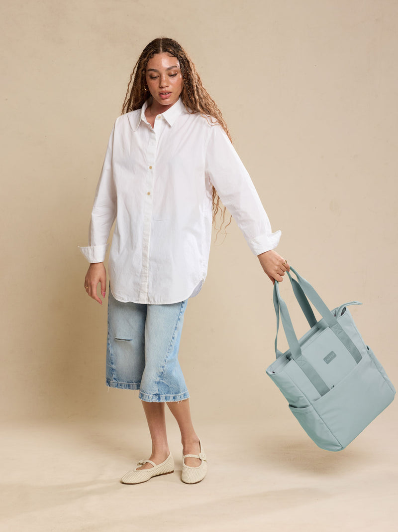 CALPAK Zippered Laptop Tote Bag in light blue Bluebell on a beige background being held by a model