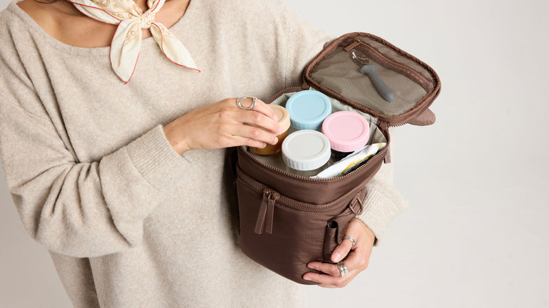 Person holding a brown CALPAK Insulated Bottle Bag with baby food containers and a spoon.