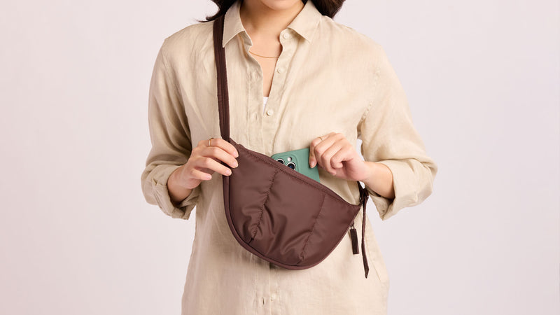 Person putting phone into the CALPAK Luka Crescent Bag in brown Walnut