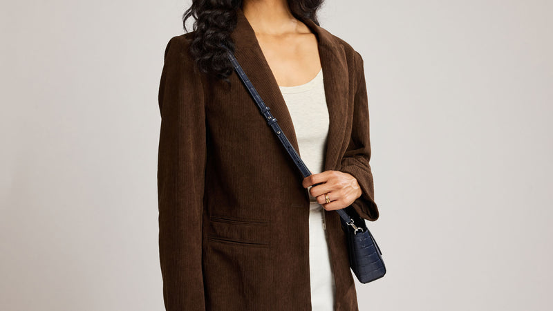 A woman wearing a brown suede-like blazer over a white top, holding a dark blue Navy Haven Mini Crossbody Tote against a plain light gray background.