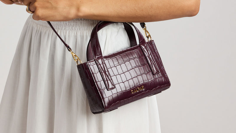 A person wearing a white dress holds a small Haven Mini Crossbody in deep red Dark Cherry in croc-embossed texture.