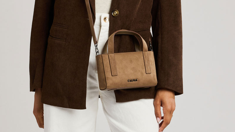 A person wearing a brown corduroy blazer and white pants carries a small Haven Mini Crossbody bag in tan Truffle Suede, against a light gray studio background.