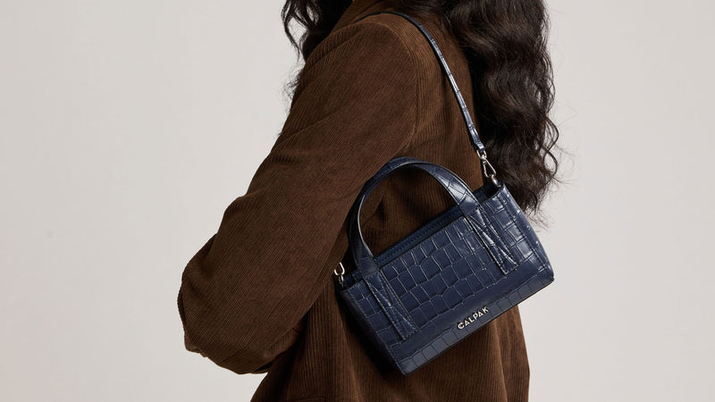 A navy blue croc-embossed Haven Mini Crossbody, shown being worn over the shoulder of a person wearing a brown corduroy jacket against a light beige background.