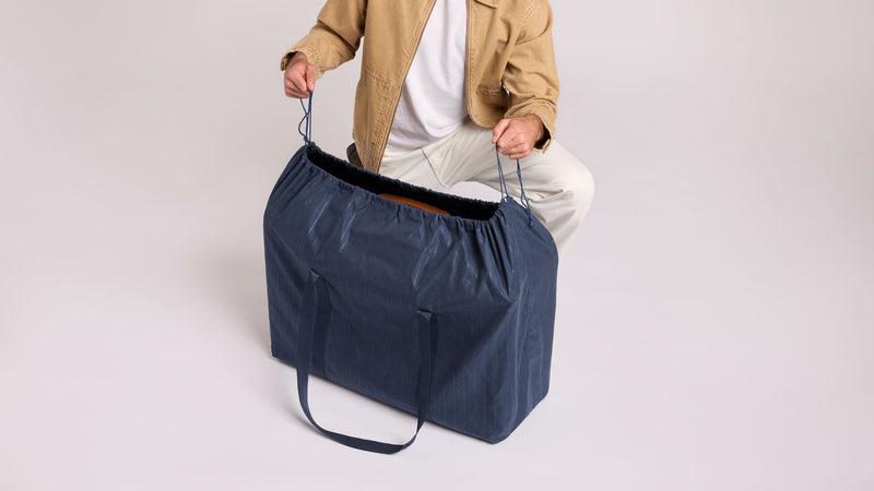 A person wearing a tan jacket, white shirt, and white pants holds the top handles of a large navy blue drawstring custom dust bag with shoulder straps that is included in the CALPAK Vault Luggage Collection, set against a plain light gray studio background.