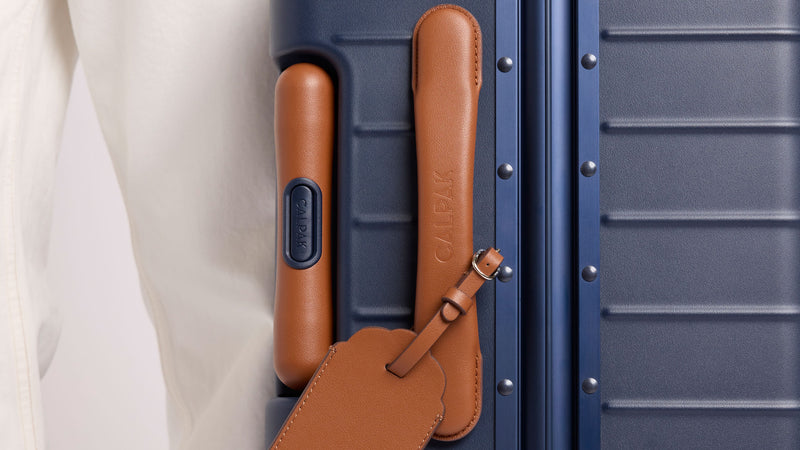 Close-up of a CALPAK Vault Luggage Navy blue hard-shell suitcase with brown leather luggage handle and matching leather luggage tag.