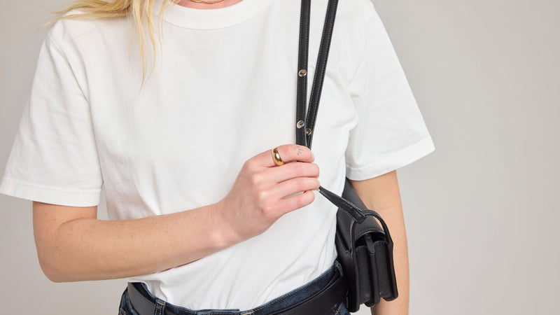 Person wearing a white t-shirt and wearing the CALPAK Canvas Colette Crossbody Bag in Black with adjustable strap on a plain background