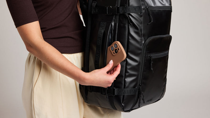 A person wearing a dark brown top and beige skirt holds a brown smartphone in their hand, showing the side pocket of the CALPAK Terra 35L backpack with a phone inside. The background is a clean, neutral gray studio backdrop.