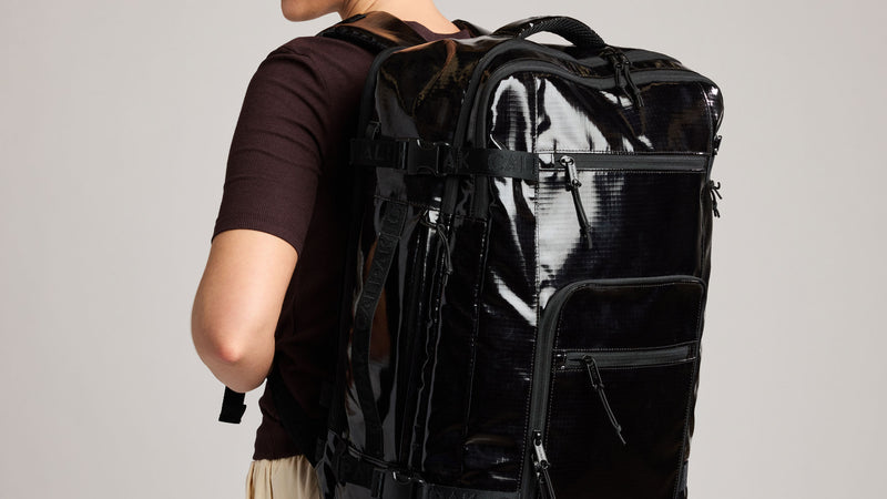 A person wearing a dark short-sleeve shirt and light-colored bottoms, shown from the back while wearing a large, glossy black CALPAK Terra 35L travel backpack with multiple compartments and straps against a plain light gray background.