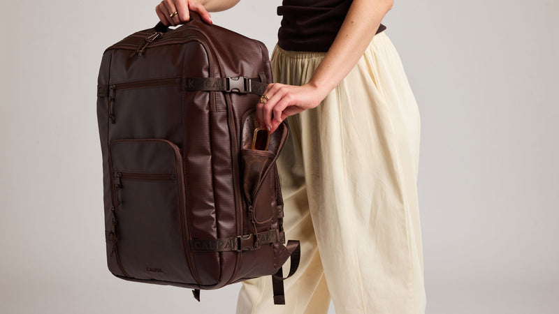 A dark brown Walnut CALPAK Terra 35L travel backpack with multiple straps and a side pocket, held by a person wearing cream-colored linen pants and a black top against a light gray studio background.