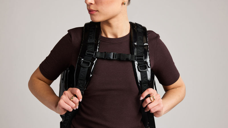 A person wearing a glossy black CALPAK Terra 35L tactical-style backpack with padded shoulder straps and front buckles, shown from the chest up against a plain light gray studio background.