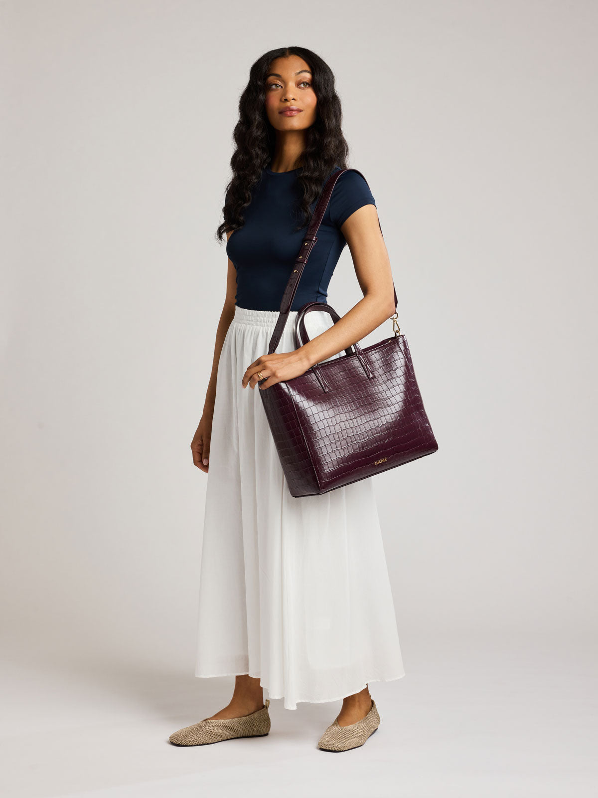 A woman modeling a dark burgundy crocodile-embossed leather shoulder bag, styled with a navy blue top, a white pleated midi skirt, and beige flats against a light gray studio background ;ATS2501-DARK-CHERRY-FAUX-CROC