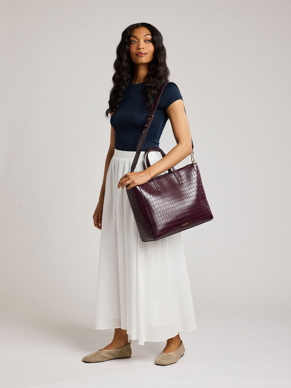 A woman modeling a dark burgundy crocodile-embossed leather shoulder bag, styled with a navy blue top, a white pleated midi skirt, and beige flats against a light gray studio background ;ATS2501-DARK-CHERRY-FAUX-CROC