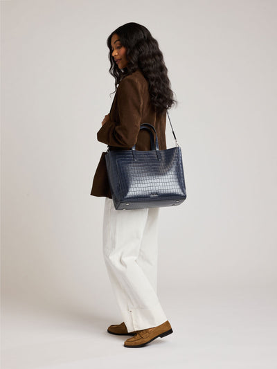 A navy CALPAK Haven 15 Laptop Tote in Navy blue crocodile-embossed with dual top handles and a matching shoulder strap against a plain white background; ATS2501-NAVY-FAUX-CROC view 2