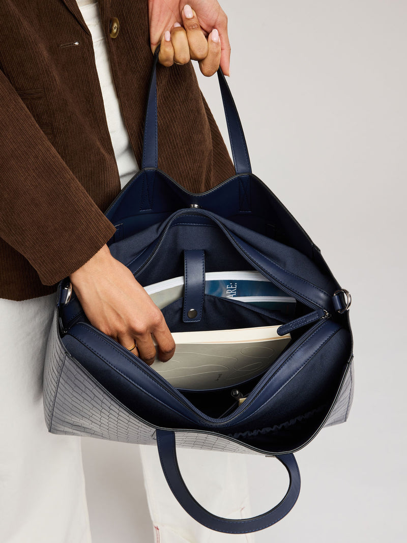 A navy blue croc-embossed tote bag with a gray plaid patterned body, shown being held by a person wearing a brown corduroy jacket