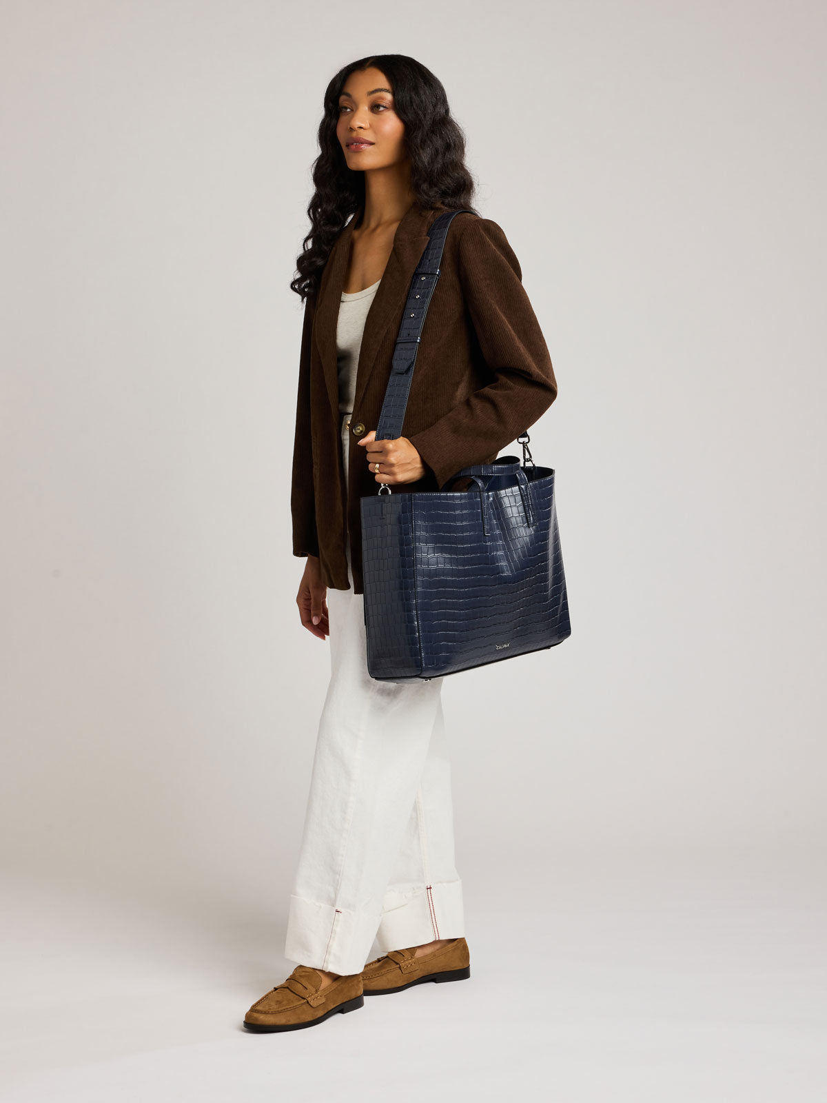A woman wearing a brown blazer, white pants, and brown loafers carries a large navy blue crocodile-embossed tote bag over her shoulder, photographed against a neutral light gray studio background; ATO2102-NAVY-FAUX-CROC