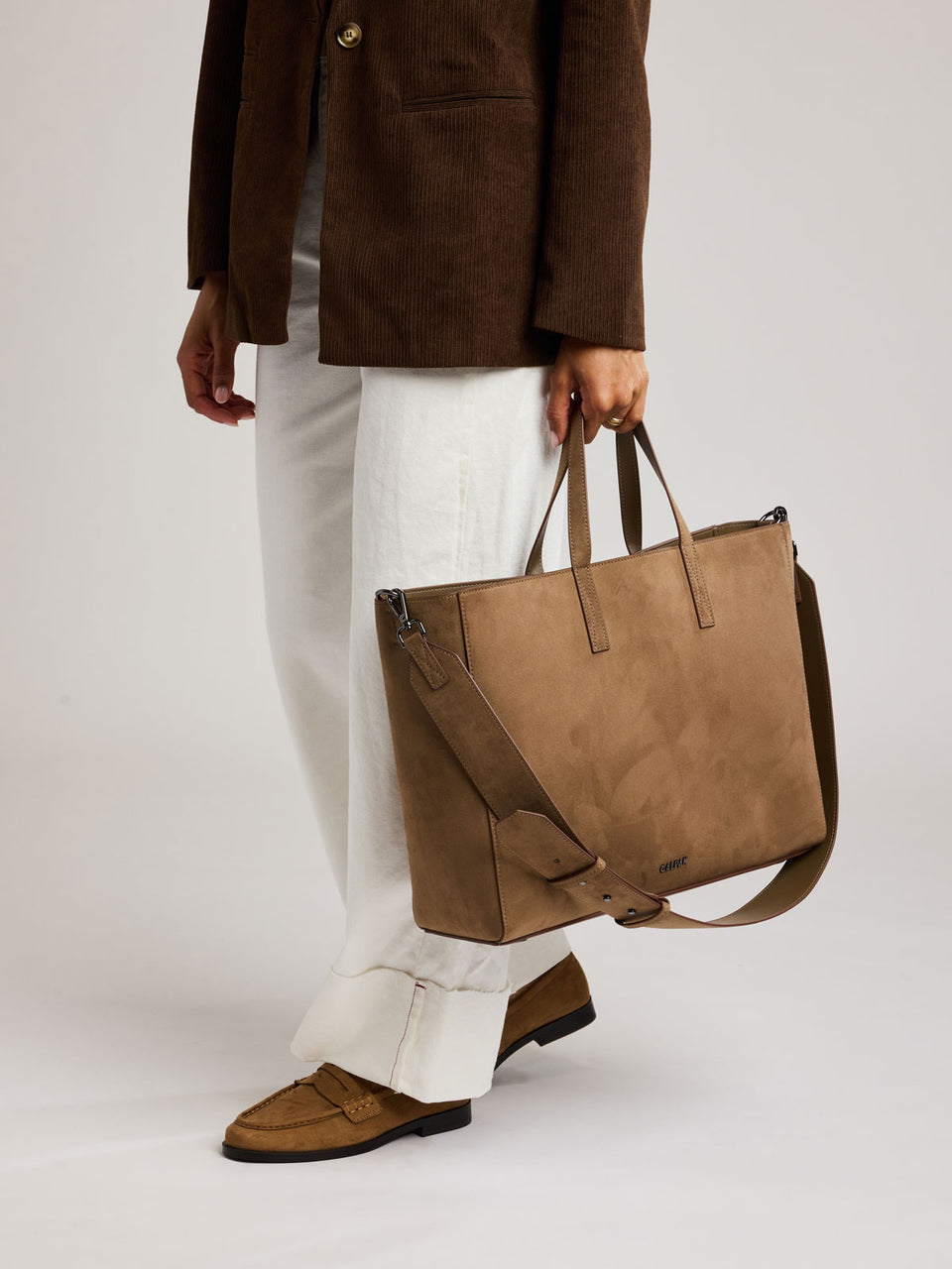 A person wearing a brown blazer, white trousers, and brown loafers carries a large tan suede tote bag with a shoulder strap against a plain light gray studio background; ATO2102-TRUFFLE-FAUX-SUEDE