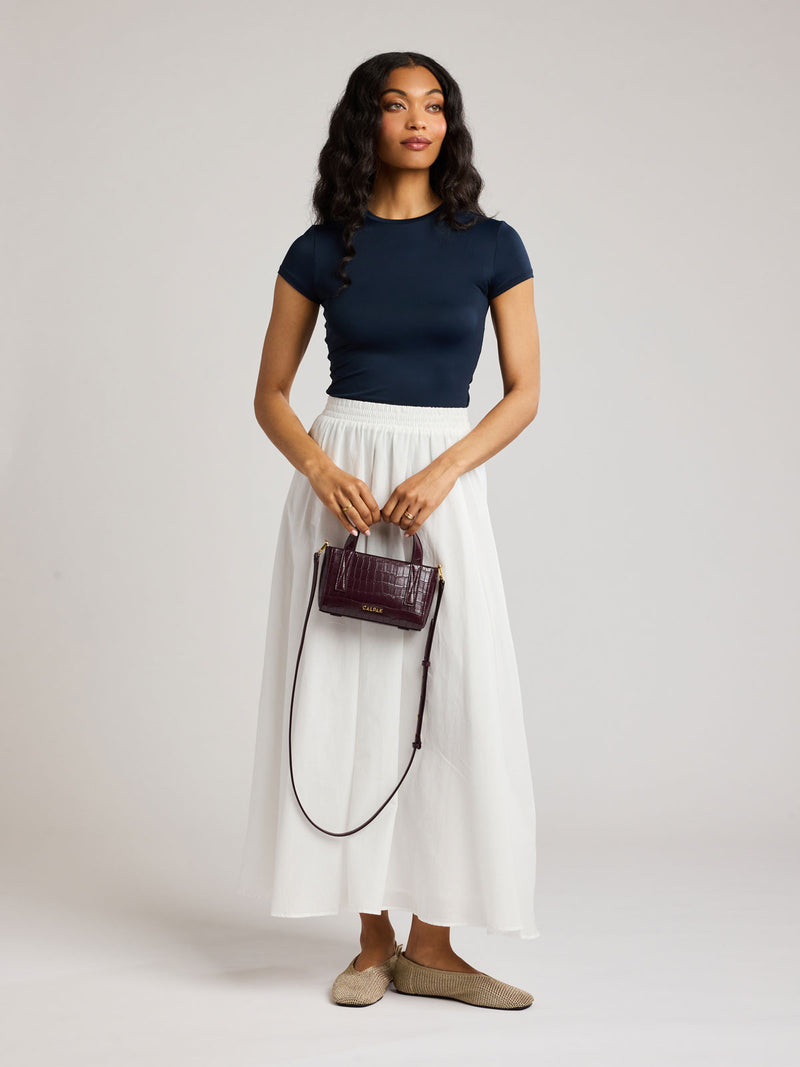 A woman modeling a navy blue short-sleeve top and a white midi skirt, holding a small Haven Mini Crossbody Bag in Dark Cherry against a light gray studio background