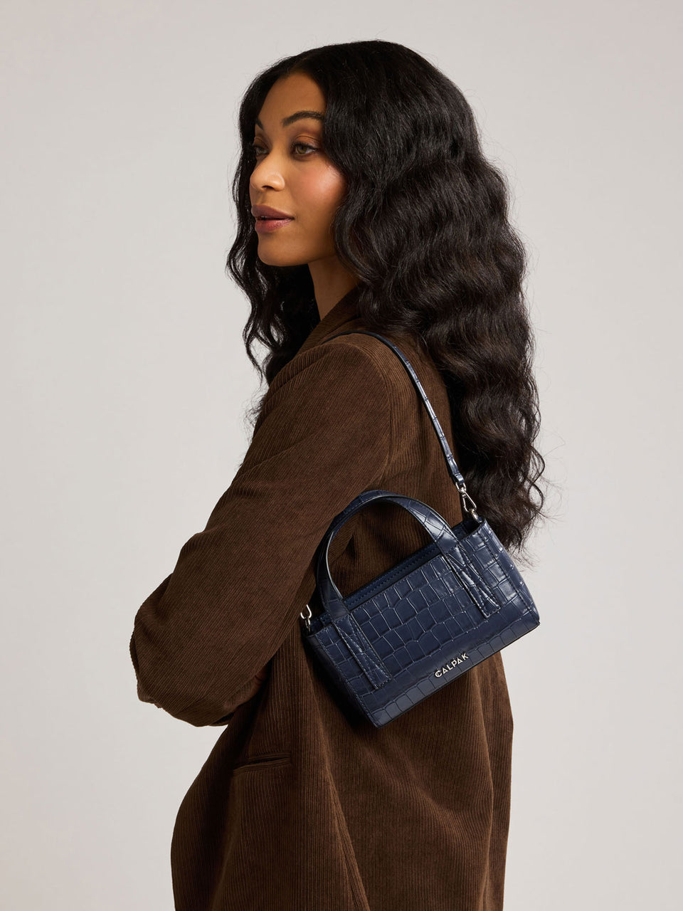 A woman with long, wavy black hair is shown in profile wearing a brown corduroy jacket and carrying a small, Mini Haven Crossbody Navy dark blue crocodile-embossed handbag; AMC2502-NAVY-FAUX-CROC
