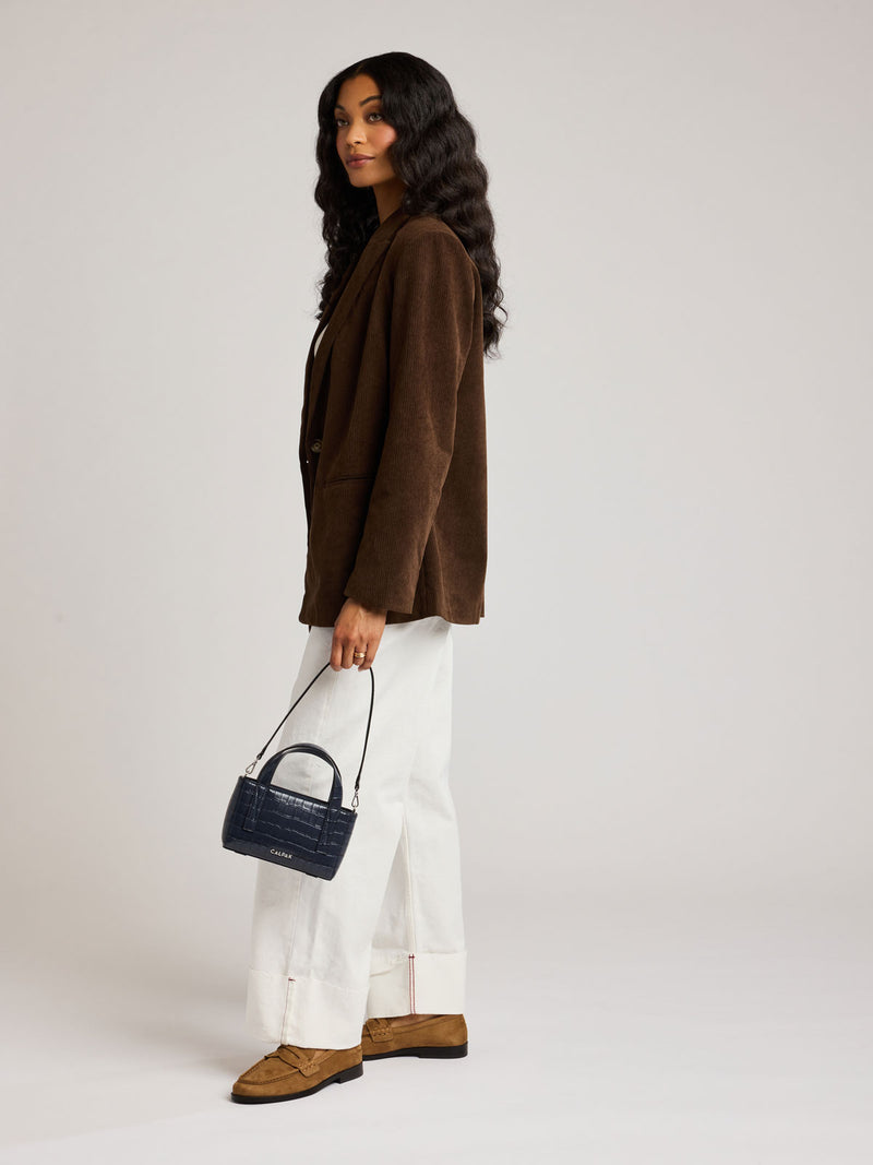 A woman stands in profile wearing a brown corduroy blazer, white wide-leg trousers, and brown suede loafers, while holding a Haven Mini Crossbody Bag in Navy dark blue croc-embossed handbag against a plain light gray studio background