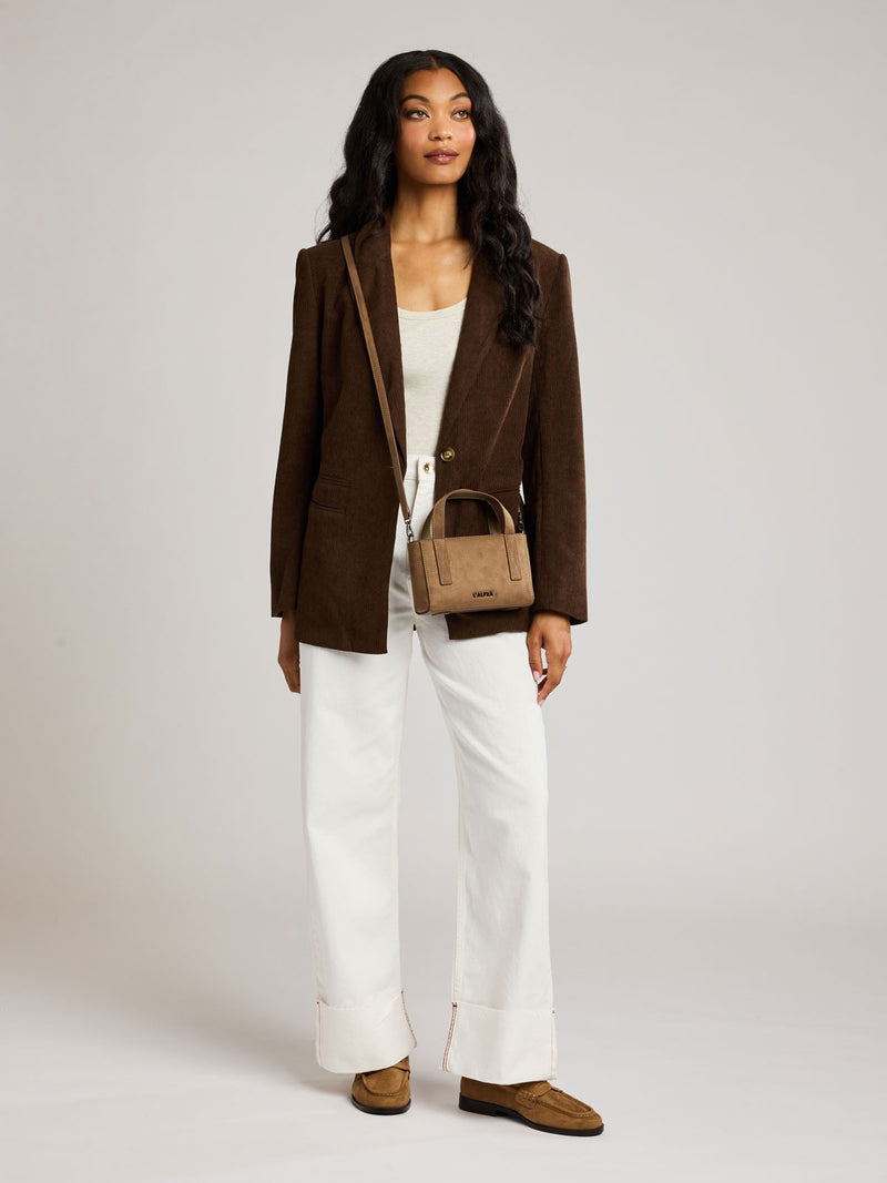 A woman stands against a light gray studio background wearing a brown corduroy blazer, white top and wide-leg white pants, with a small Haven Mini Crossbody Bag in tan brown Truffle Suede
