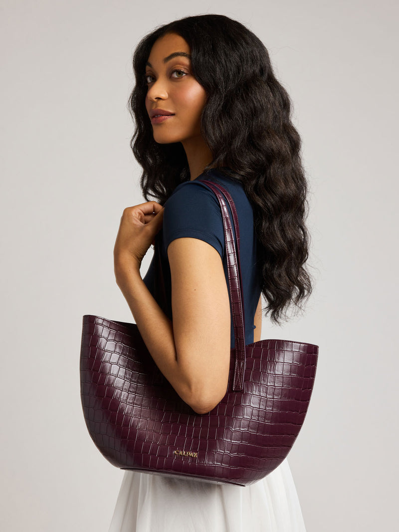 A woman with long, wavy dark hair is shown from the side/back, wearing a Haven Tulip Tote in Dark Cherry that is crocodile-embossed over her shoulder against a neutral light gray studio background