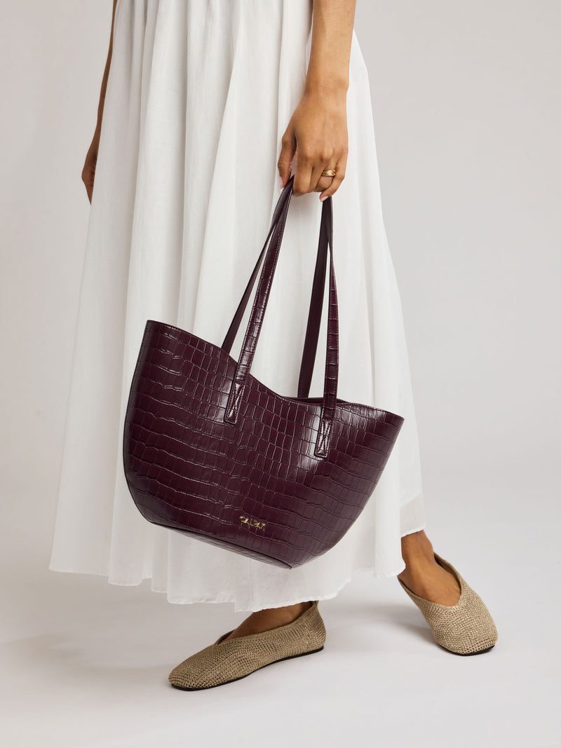 A person wearing a white maxi dress and beige flat shoes holds a large, Haven Tulip Tote in Dark Cherry in crocodile-embossed tote bag with double shoulder straps