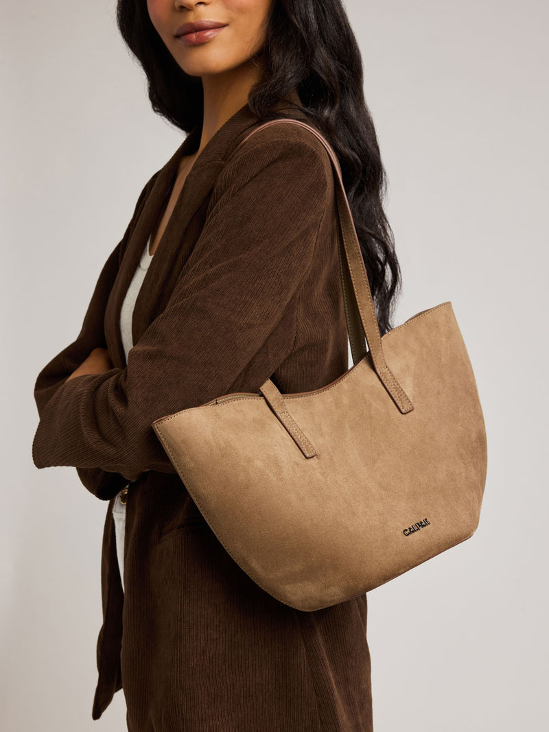 A woman wearing a brown corduroy cardigan carries a Haven Tulip Tote bag in brown tan Truffle Suede with a set against a plain light gray studio background