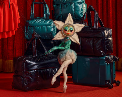 A styled studio scene with multiple shiny metallic teal travel bags and a suitcase arranged against a red curtain backdrop, with a flower-doll figure seated in front. No person or real environment is present; the doll is a prop, so this is Product/Packaging rather than Lifestyle.