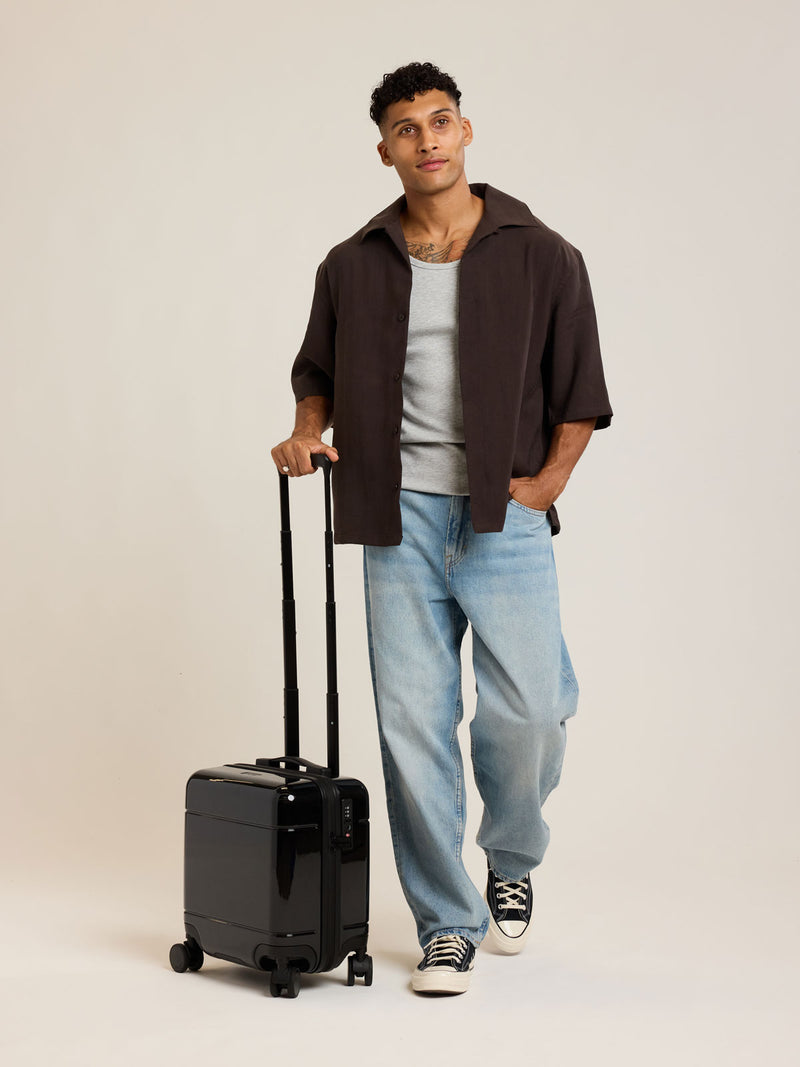 Model with CALPAK Glazed Black Hue Mini Carry On suitcase with a black handle on a white background