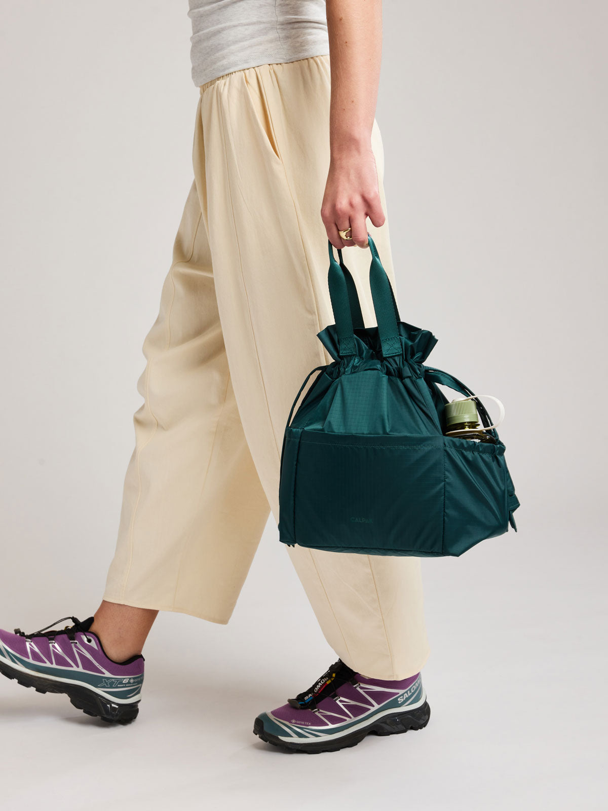 A person wearing cream wide-leg pants and purple trail running shoes carries a dark teal green Kale CALPAK Insulated Lunch Bag with top handles, shown against a plain white background; ALB2001-KALE