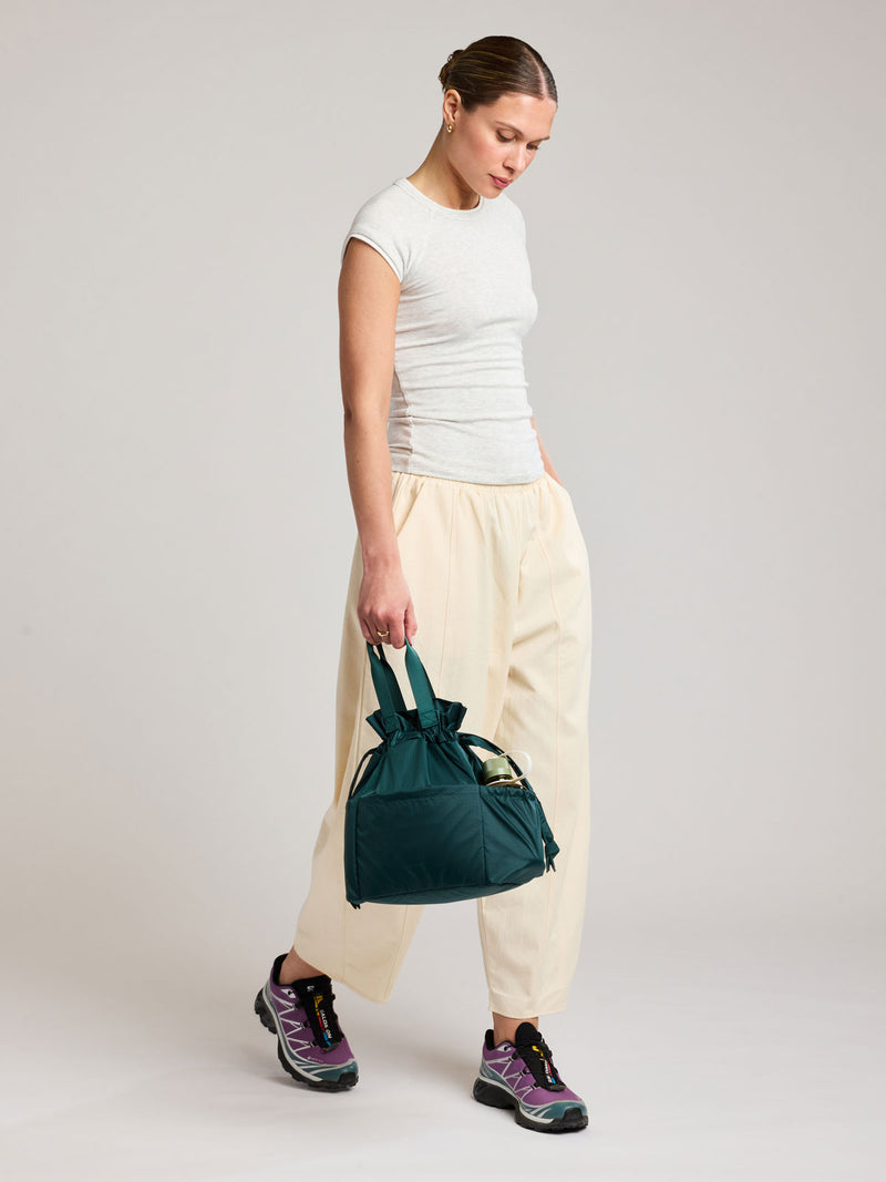 A woman stands in a studio wearing a white fitted short-sleeve top, cream drawstring pants, and purple athletic shoes, while holding a dark teal green Kale CALPAK Insulated Lunch Bag with top handles, shown against a plain white background