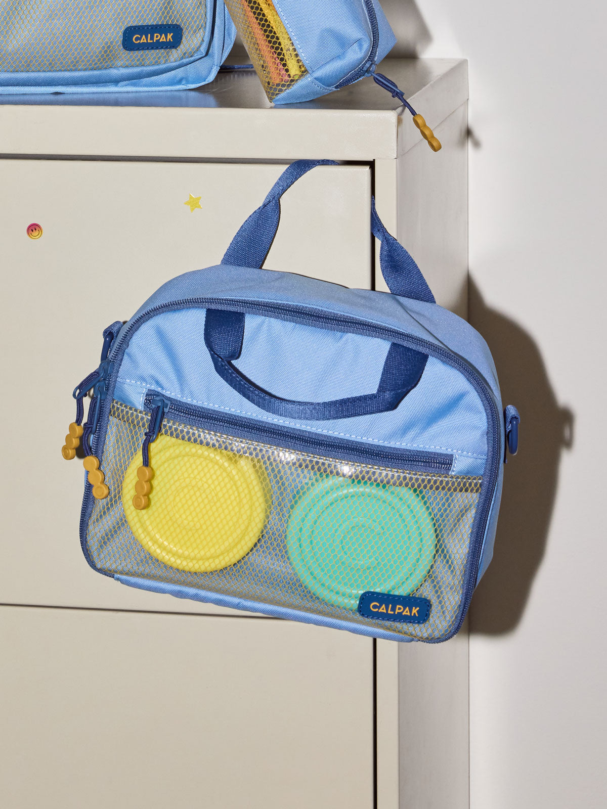 A CALPAK Kids Lunch Bag in Blue with a top handle and zipper, shown hanging on a white cabinet; CKLB2501-BLUE