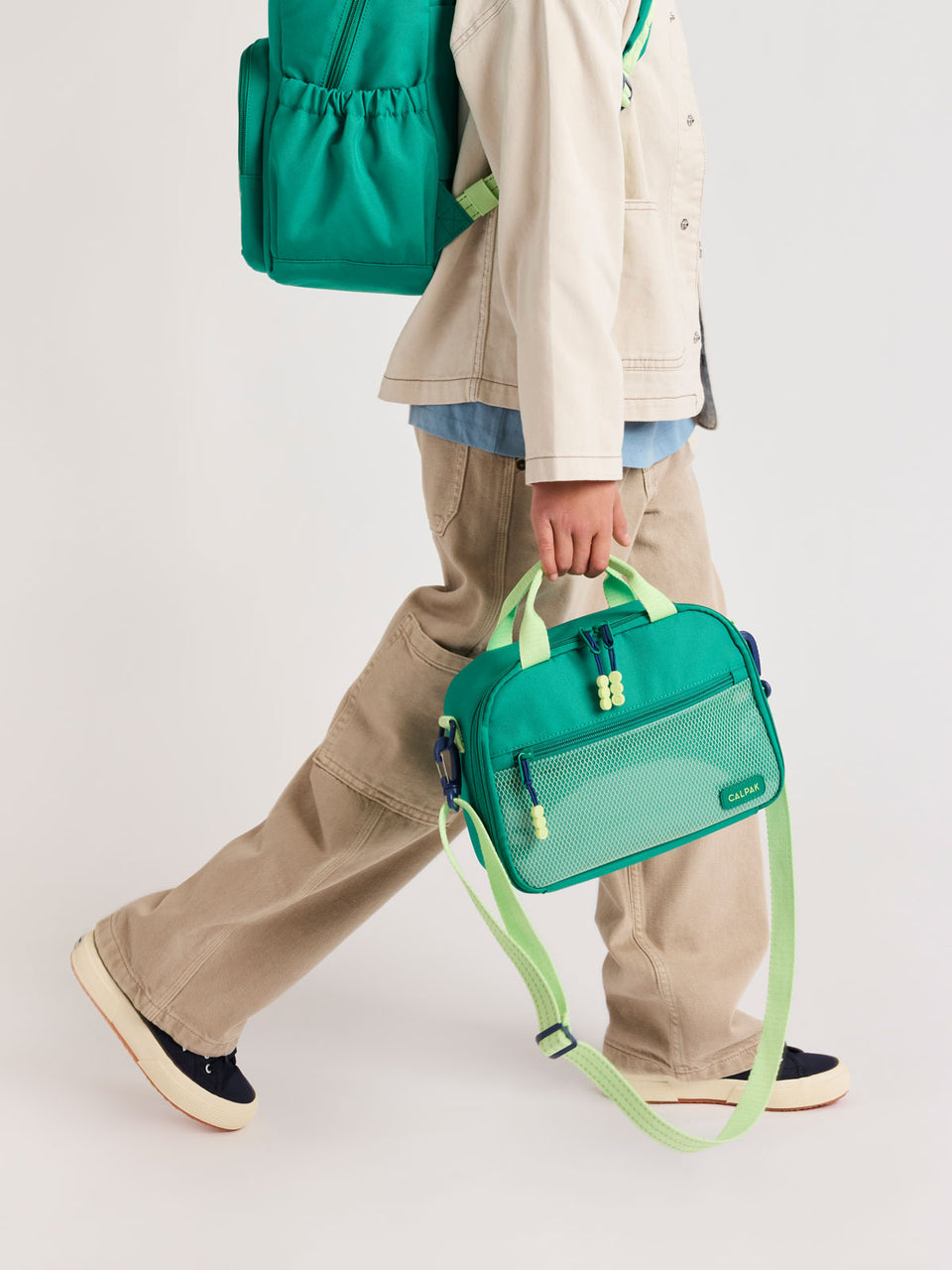 Person holding the Green CALPAK Kids Lunch Bag with lime green accents and front mesh pocket on a white background; CKLB2501-GREEN