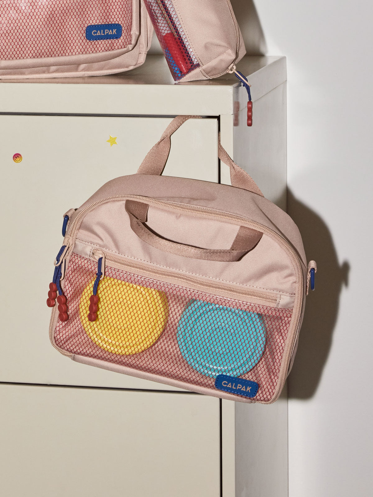 A CALPAK Kids Lunch Bag in Khaki with a top handle and zipper, shown hanging on a white cabinet; CKLB2501-KHAKI