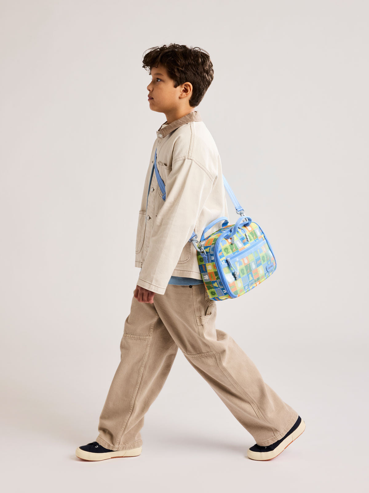 Boy wearing the crossbody of CALPAK Kids Lunch Bag with Miffy and animal pattern with mesh front pocket and top handles on a white background; CKLB2501-MIFFY-BLUE-ZOO-TRIP