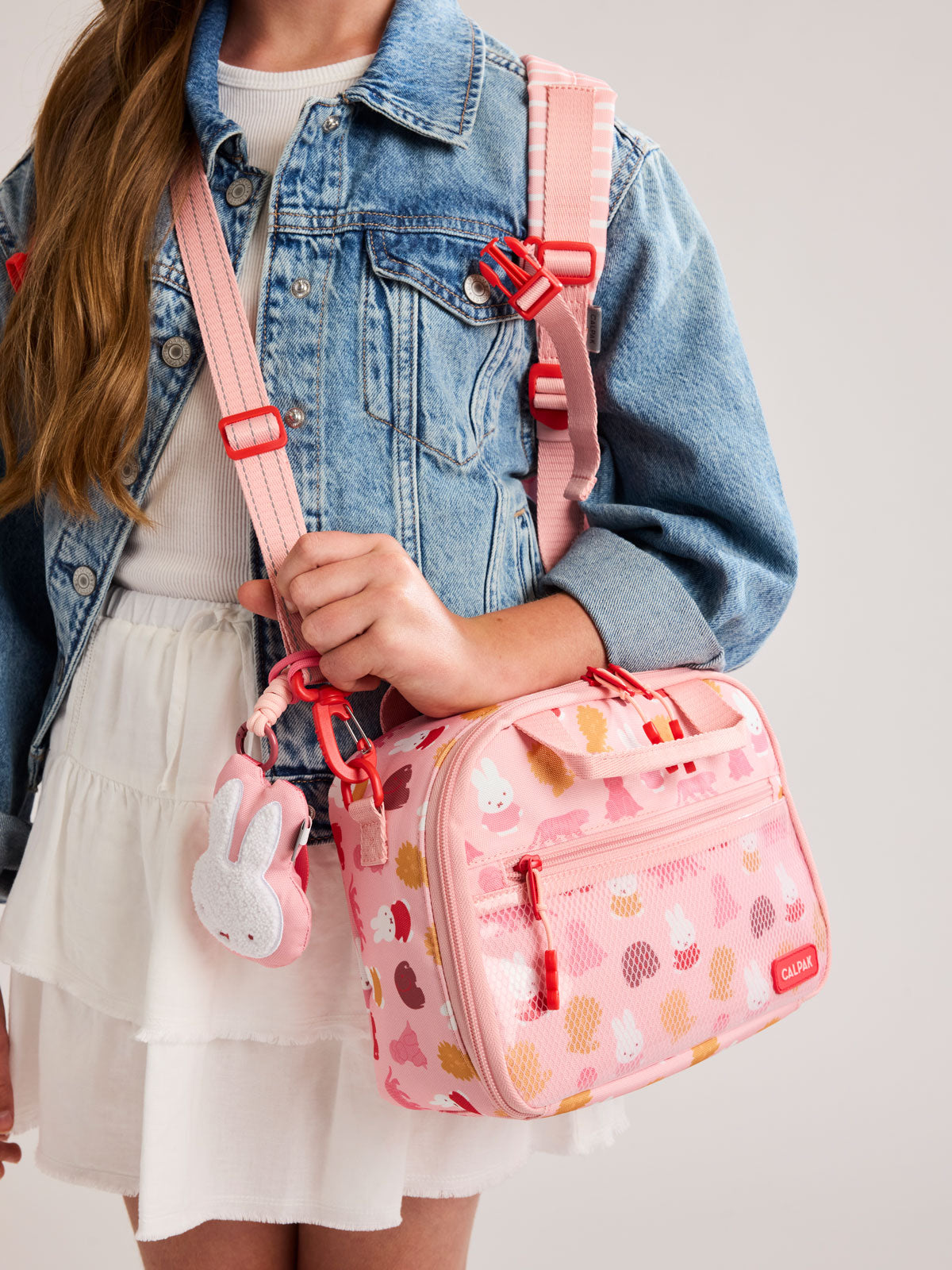 Girl wearing the Pink CALPAK Kids Lunch Bag with Miffy and animal pattern with mesh front pocket and top handles on a white background; CKLB2501-MIFFY-PINK-ZOO-TRIP