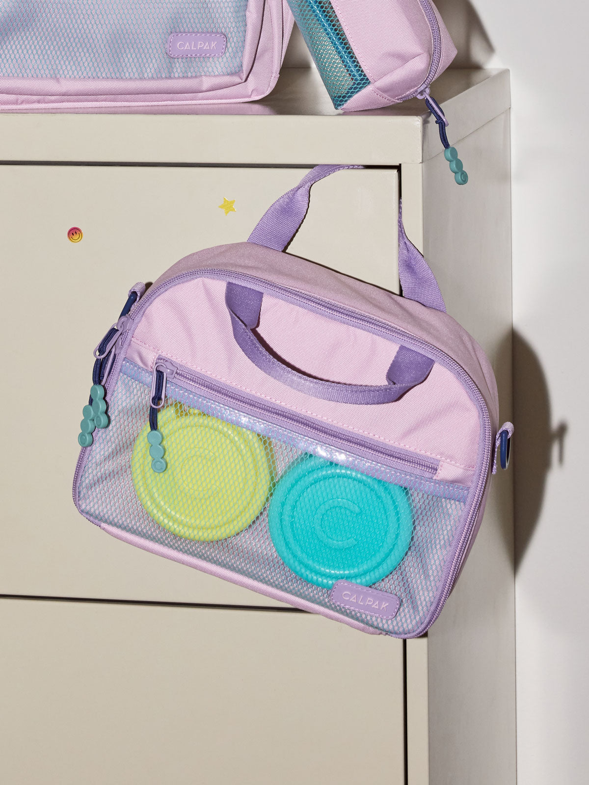 A CALPAK Kids Lunch Bag in Orchid Pink with a top handle and zipper, shown hanging on a white cabinet; CKLB2501-ORCHID-PINK