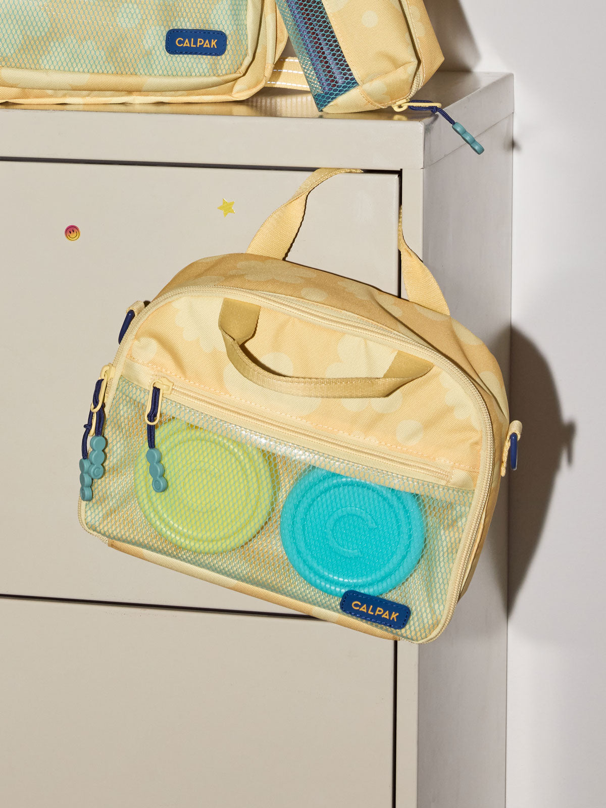 A CALPAK Kids Lunch Bag in yellow bloom  with a top handle and zipper, shown hanging on a white cabinet; CKLB2501-YELLOW-BLOOM
