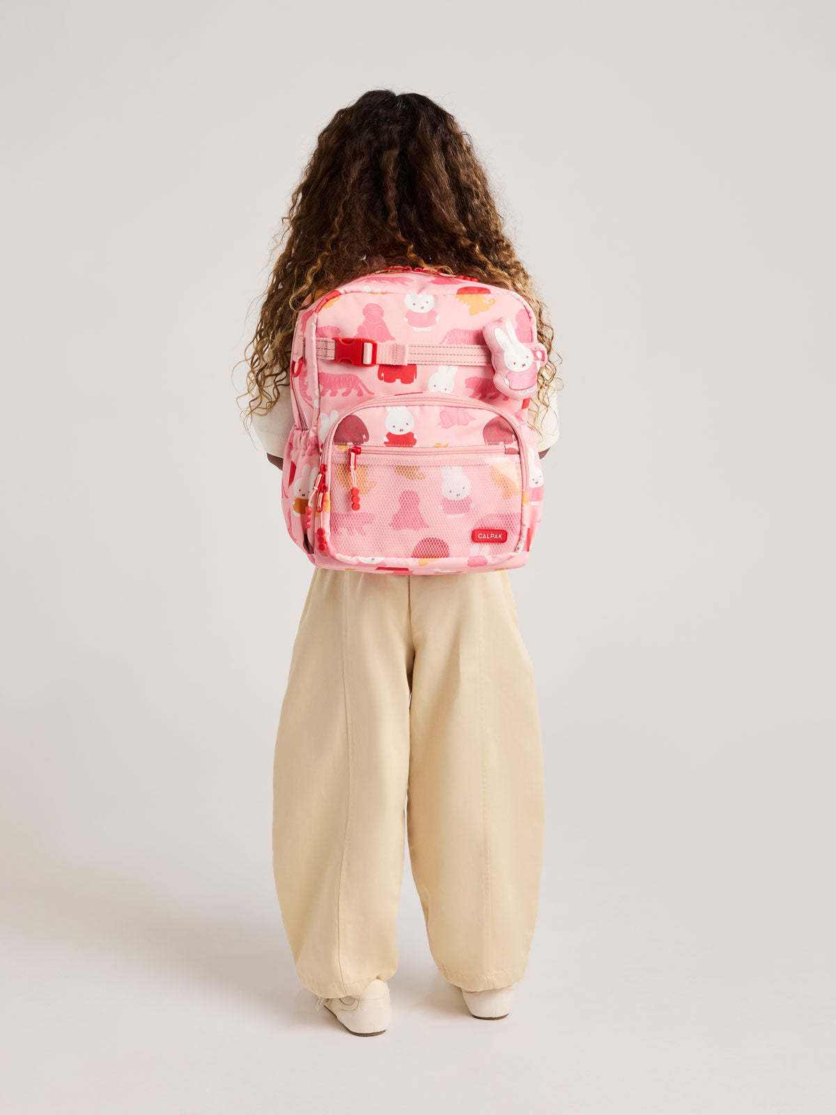Person wearing a CALPAK Kids Medium Backpack with Miffy and pink colorful animal pattern on a white background; CKBM2501-MIFFY-PINK-ZOO-TRIP