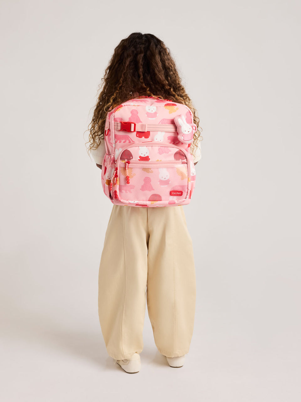 Person wearing a CALPAK Kids Medium Backpack with Miffy and pink colorful animal pattern on a white background; CKBM2501-MIFFY-PINK-ZOO-TRIP