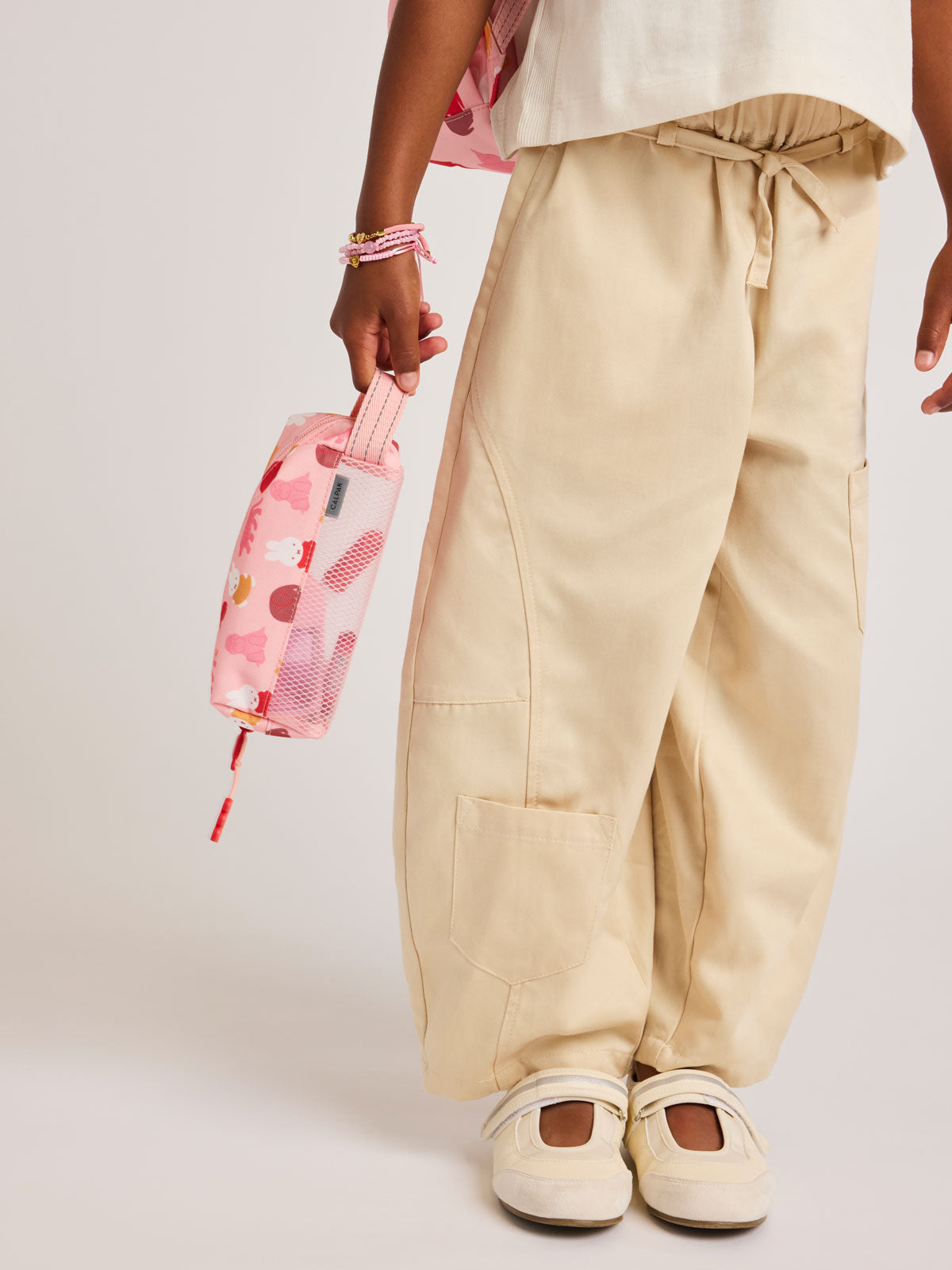 Person wearing beige pants holding a pink Colorful CALPAK Kids Pencil Case with Miffy and cartoon designs on a white background in Miffy Pink Zoo Trip; CKPC2501-MIFFY-PINK-ZOO-TRIP