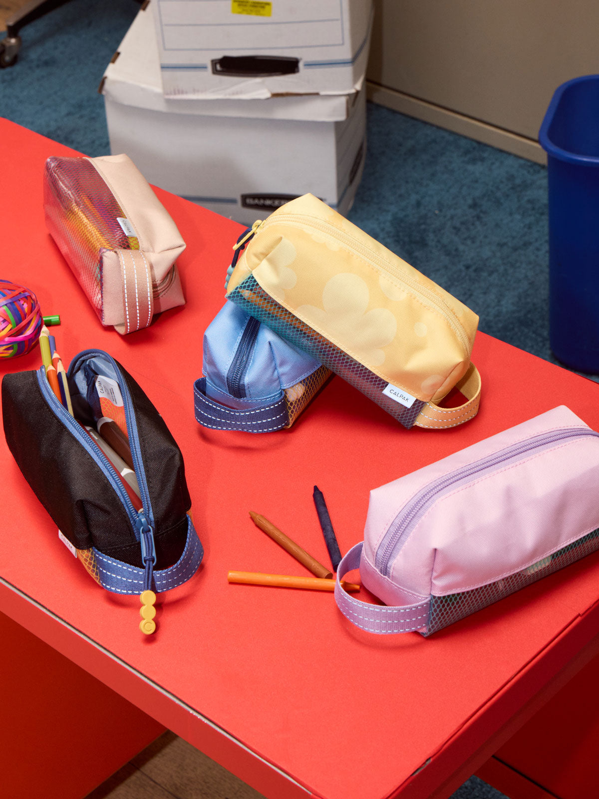 A collection of CALPAK Kids Pencil cases pencil cases arranged on a bright red table, with pens placed in front of them; CKPC2501-YELLOW-BLOOM