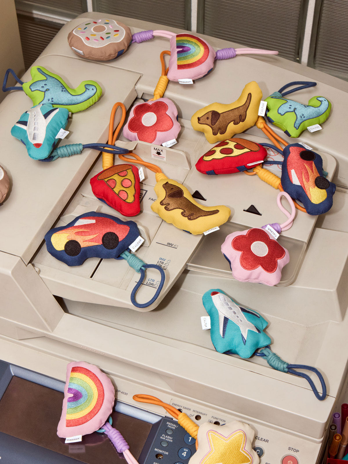A collection of colorful CALPAK Pluffy Bag Charms shaped like cars, flowers, dinosaurs, a pizza slice, a rocket ship, and rainbows, arranged on top of a beige photocopier; APLF2501-DOG